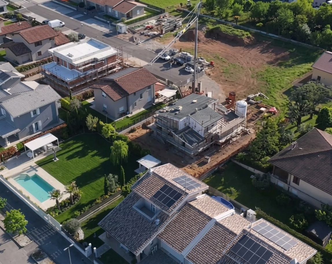 Vista aerea più ampia del quartiere attorno al cantiere, con case, giardini e campi aperti oltre.